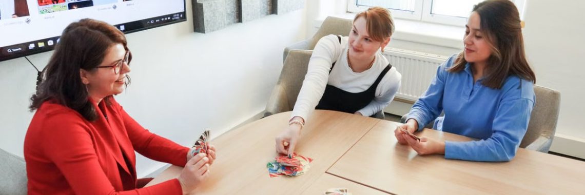 Three women enjoying a card game at a wooden table in a modern office setting.